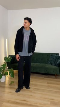 Black fleece zip-up jacket over a gray t-shirt with pink text, paired with loose black pants and black shoes; indoor setting.