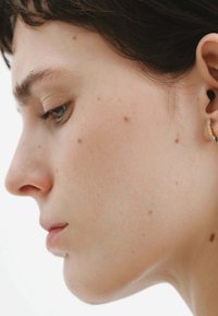 Vista de perfil de una persona con cabello corto y oscuro, piel suave y pequeñas manchas en la cara. Se puede ver un pendiente de oro y plata en la oreja.