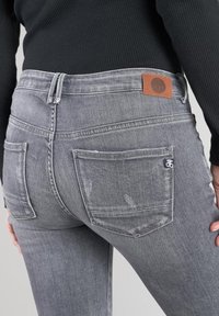 Jean en denim gris au design ajusté, texture usée, et deux poches arrières. Présente une patch en cuir brun au niveau de la ceinture.