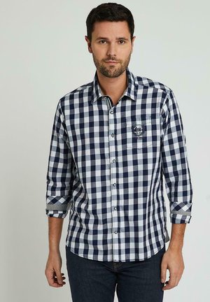 Chemise à carreaux bleu marine et blanche à manches longues, fermée par boutons à l'avant, avec une poche poitrine arborant un logo. Fabriquée en tissu tissé.