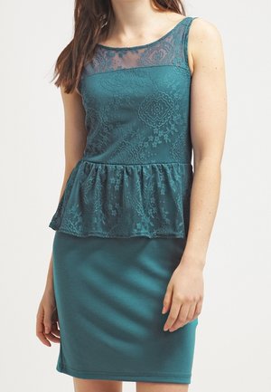 Kobieta w turkusowej sukience bez rękawów z koronkowym gorsetem, przezroczystym dekoltem i detalem peplum w talii na jednolitym tle.