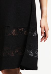 Černá texturovaná sukně s dvěma vodorovnými průhlednými krajkovými panely blízko lemu, které ukazují květinové vzory, s viditelnou pravou rukou vedle ní.
