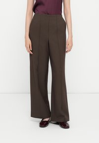 Pantaloni ampi marroni con una superficie liscia, caratterizzati da due pieghe frontali e una vita alta, abbinati a una maglietta viola scuro e scarpe lucide.