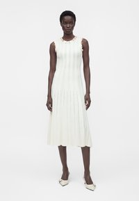 Femme debout dans une robe midi en maille blanche sans manches avec une bordure florale aux épaules et des talons blancs à bout pointu.