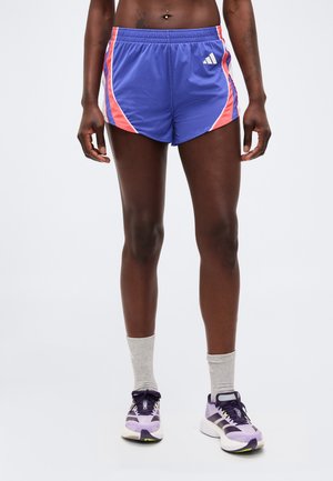 Athlète portant un short de course bleu et rose, des chaussettes grises montantes et des chaussures de course violettes, debout sur un fond uni.