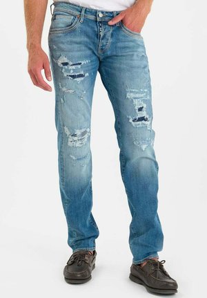 Uomo che indossa jeans azzurri strappati con toppe, camicia bianca e scarpe casual marrone scuro, in piedi con una mano in tasca.