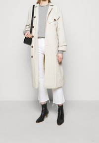 Gabardina beige con botones marrones, combinada con un suéter gris y jeans blancos cortos, complementada con botines negros.