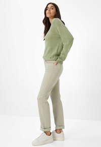 Donna in piedi di lato che indossa un maglione verde chiaro, pantaloni beige con risvolti e scarpe da ginnastica bianche su uno sfondo bianco semplice.