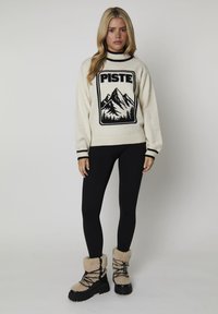 Crème sweater met een graphic van een zwarte berg en de tekst "PISTE", gecombineerd met zwarte leggings en tan, met bont gevoerde laarzen met zwarte zolen.