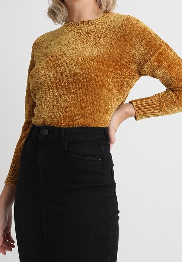 Femme portant un pull texturé jaune moutarde glissé dans une jupe en denim noire taille haute, sa main reposant sur sa hanche.