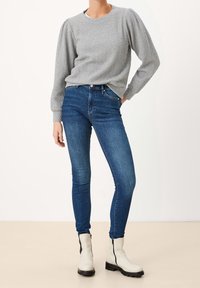 Grau strukturierter Pullover mit langen Ärmeln, kombiniert mit dunkelblauen Skinny Jeans und weißen Stiefeln mit schwarzen seitlichen Reißverschlüssen.