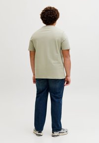 Άνδρας με σγουρά μαλλιά στέκεται με την πλάτη, φορώντας ανοιχτοπράσινο κοντομάνικο πουκάμισο, σκούρο μπλε τζιν και λευκά αθλητικά παπούτσια σε απλό φόντο.