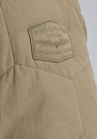 Chaqueta de pana caqui con una superficie texturizada y un parche cosido que presenta "PME Legend Aviator" y un detalle de emblema alado.