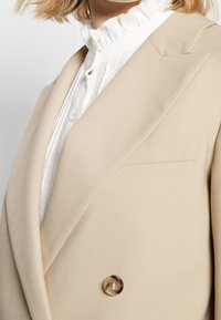 Nahaufnahme einer Person, die ein beiges Blazer über einer weißen Rüschbluse trägt, wobei der Revers, der Knopf und der obere Brustbereich sichtbar sind.