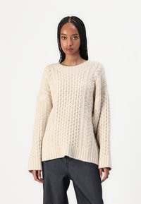 Cremefarbener Strickpullover mit strukturiertem Muster, langen Ärmeln und rundem Halsausschnitt. Der Schlitz endet auf Taillenhöhe.