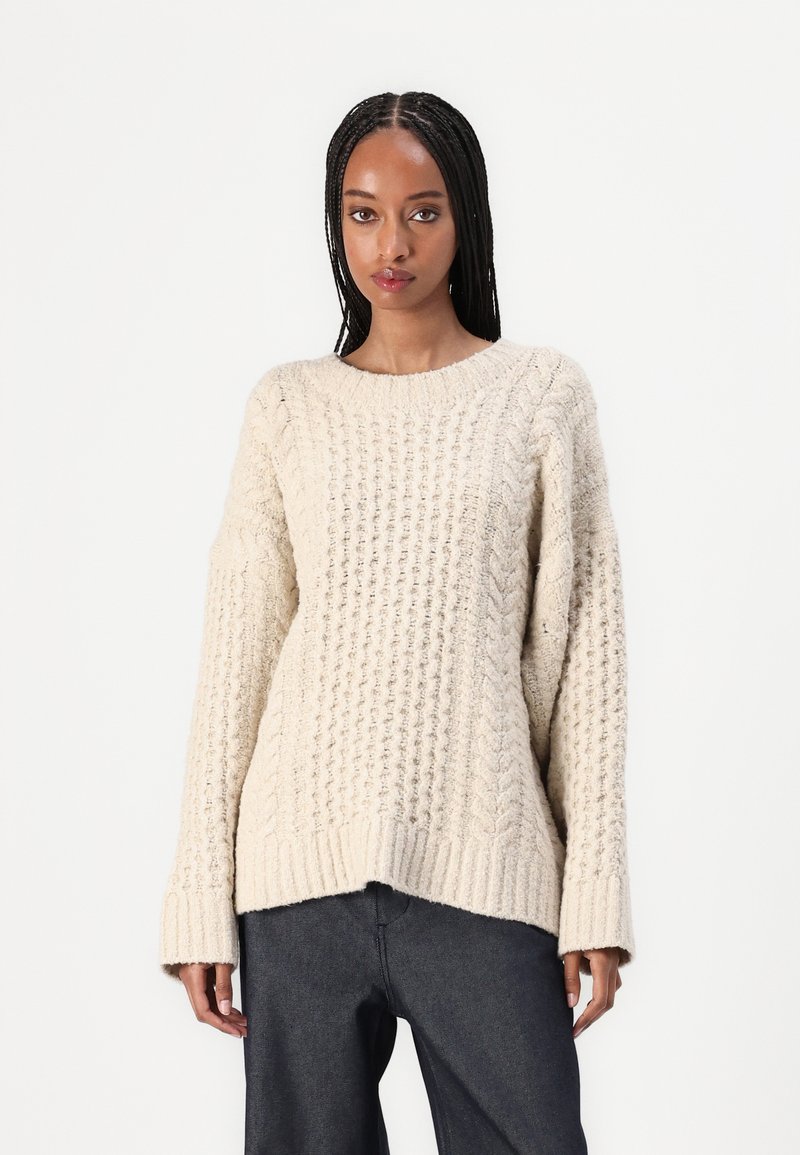 Cremefarbener Strickpullover mit strukturiertem Muster, langen Ärmeln und rundem Halsausschnitt. Der Schlitz endet auf Taillenhöhe.