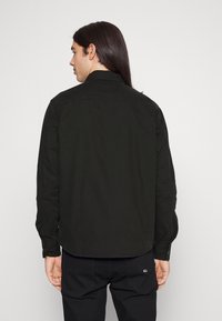 Camisa negra de manga larga con un cuello tipo camisa, detalle de costura en el yugo trasero y puños abotonados, confeccionada en una tela suave con un acabado mate.