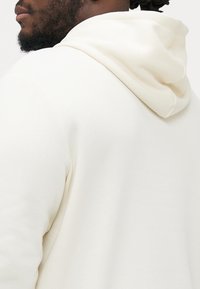 Off-white Hoodie aus weichem Stoff, mit einer geräumigen, runden Kapuze und einer glatten Textur. Sichtbare Nähte entlang der Säume.