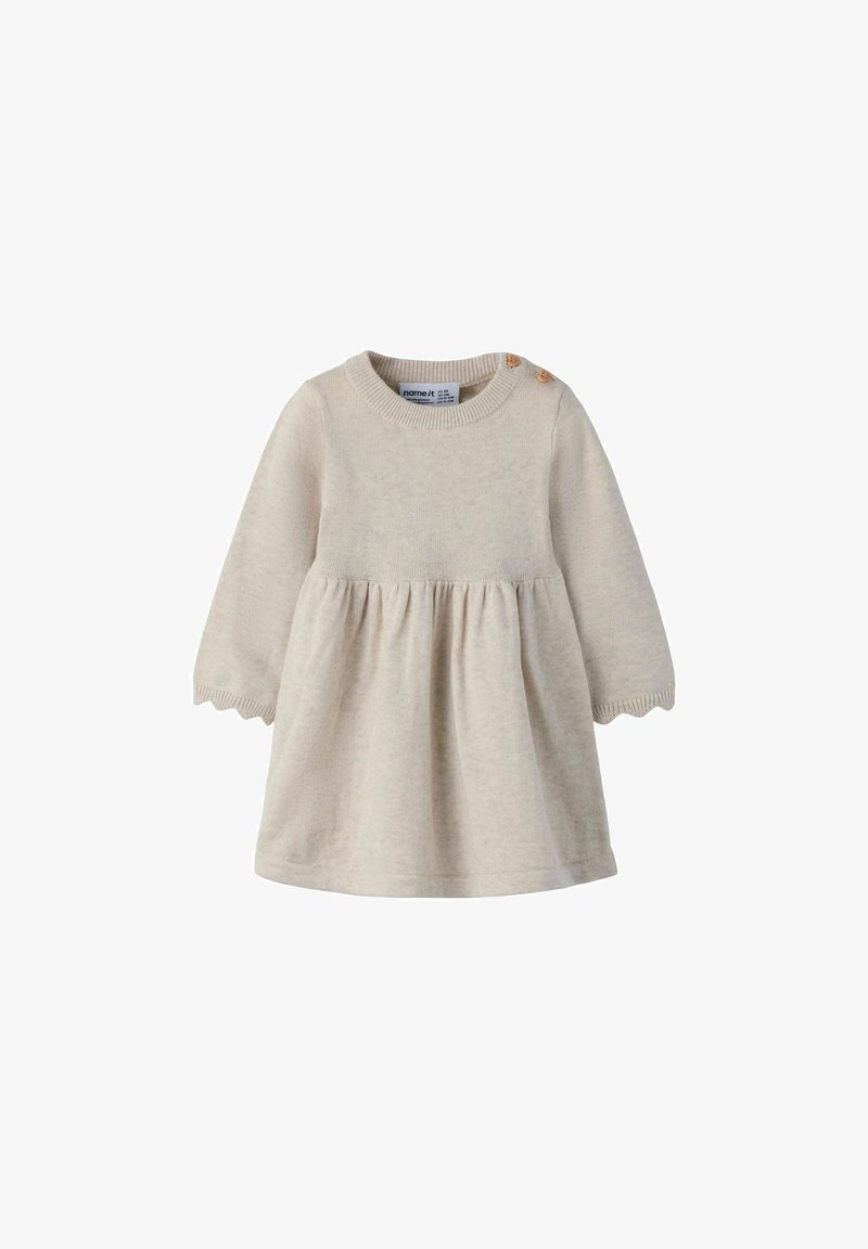Robe bébé longue beige en tricot avec deux boutons en bois sur l’épaule et bords festonnés aux poignets.