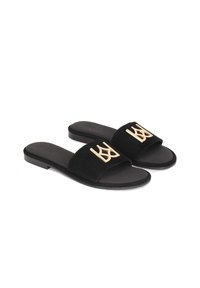 Crne suede natikače s ravnim đonom imaju zlatni metalni logotip na remenu. Jednostavan dizajn s otvorenim nožnim prstima.