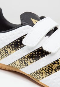 Zapatilla deportiva blanca con acentos de rayas negras y doradas. Presenta correas de velcro y una superficie texturizada. Suela de goma con detalles en patrones.