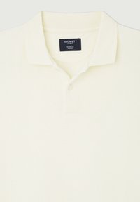 Pikétröja i ljus krämfärgad, strukturerad tyg. Har en klassisk krage, tre knappar fram och en etikett med "Hackett"-logotyp.