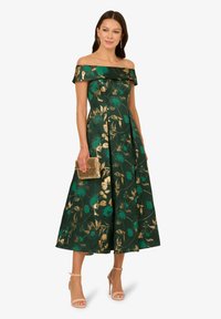 Zelené květinové šaty bez ramínek se zlatými akcenty, A-line sukně a texturovaná látka. V kombinaci se zlatým psaníčkem a sandály na podpatku.