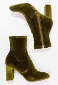Bottines en velours vert olive avec des talons carrés et des zips latéraux, présentées en vue latérale et inférieure sur un fond blanc.
