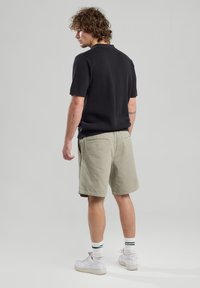 Musta lyhythihainen poolopaita, vaaleanvihreät shortsit, valkoiset sneakerit vihreillä yksityiskohdilla, seisoo selkä kameraan päin, tekstuurinen kangas.