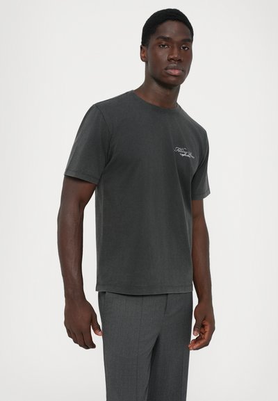 Adult man wearing dark gray short-sleeve T-shirt with small white logo on chest and matching gray trousers, standing against white background.