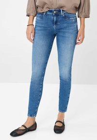 Jeans skinny bleu avec un délavage, design à cinq poches et fermeture à bouton métallique, associés à des chaussures plates en mesh noires.