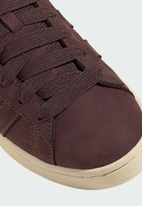 Brown suede sneaker with textured fabric laces, featuring a rounded toe and beige rubber sole. Visible stitching accents the design.