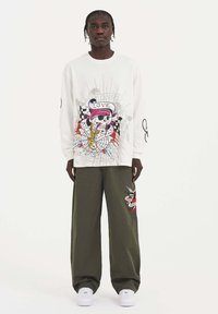 Witte lange mouwen shirt met een kleurrafische afbeelding van een schedel en tekst, gecombineerd met losse olijfgroene broek versierd met borduursel en een patch.
