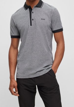 Homme portant un polo gris avec col et bords de manches noirs, logo "BOSS" brodé sur la poitrine, et pantalon noir, une main dans la poche.