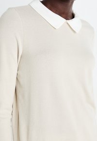 Beige gebreide trui met een witte, button-down kraag. Heeft een zachte textuur en een aansluitend ontwerp, met de nadruk op de romp en schouderpartij.