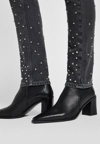 Botas de tornozelo pretas em couro com bico pontiagudo e salto bloco, combinadas com jeans cinzas adornados com tachas prateadas por toda a parte.