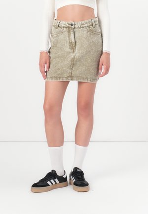 Khaki džínová sukně s přiléhavým střihem, dvěma předními kapsami a poutky na pásek. Nosit s černými teniskami a bílými ponožkami. Hladká textura.