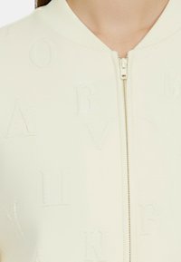 Giacca bomber color crema chiaro con chiusura a zip, tessuto testurizzato con lettere in rilievo e colletto e polsini a costine.
