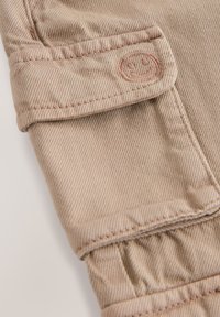 Beige stof met een subtiele diagonale textuur, voorzien van een opgehælde boord en geborduurd smiley-logo. Schone stiksels en een zachte afwerking.