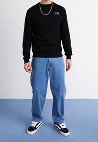 Felpa nera con logo, jeans blu e sneakers bianco e nere. Il materiale sembra morbido; la felpa ha polsini e orlo a costine.