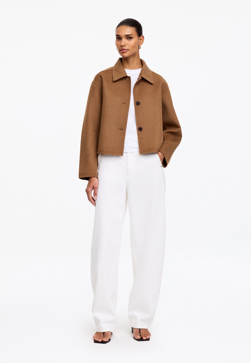 Femme portant une veste marron boutonnée, un pantalon blanc à jambes larges, une chemise blanche et des talons ouverts noirs, debout avec une main dans la poche.