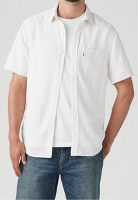 Chemise à manches courtes en tissu blanc avec des fines rayures bleues et rouges, dotée d'une fermeture à boutons sur le devant et d'une poche poitrine. Portée sur un T-shirt blanc.