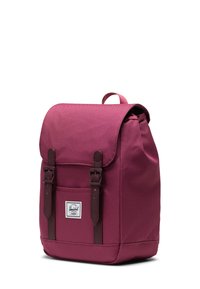 Bordeauxfärgad tygryggsäck med klaffstängning, bruna läderremmar, framficka och en logolapp. Slät textur och rektangulär form.