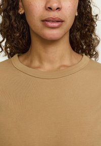 Bežs, īsām piedurknēm kokvilnas tops ar apaļu kakla izgriezumu un gludu tekstūru, ar smalkām šuvju detaļām ap apkakli.