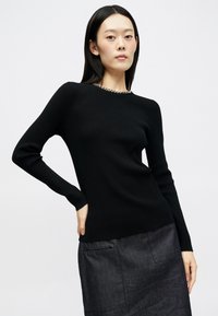 Μαύρο ριμπ πουλόβερ με μακριά μανίκια και στρογγυλή λαιμόκοψη, με διακοσμητική αλυσίδα στη λαιμόκοψη. Φορεμένο με σκουρόχρωμη φούστα.