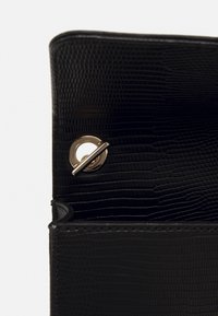 Černá texturovaná kabelka s klopou otevřenou, která odhaluje zlatý kruhový zacvakávací uzávěr a embosovaný vzor ještěra na hladkém koženém povrchu.