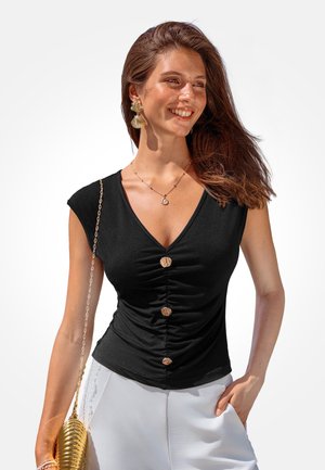 Lächelnde Frau mit langen braunen Haaren trägt ein schwarzes gerafftes ärmelloses Oberteil mit großen Knöpfen, weiße Hose, goldene Ohrringe, Halskette und goldene Clutch-Tasche.