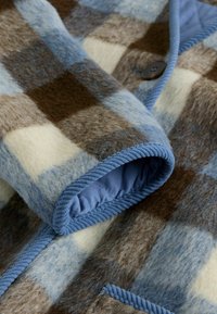 Wełniany płaszcz w kratę z niebieskimi, brązowymi i kremowymi kwadratami. Miękka faktura z niebieską podszewką i pasującymi detalami w formie lamówek. Zapięcie na jeden guzik.