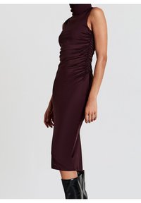 Femme portant une robe midi moulante sans manches à col montant de couleur violet foncé avec des côtés froncés, associée à des bottes noires brillantes montant jusqu'aux genoux, debout de profil.