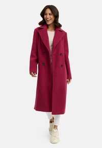 Fuchsia ullkappa med dubbelknäppt design, stor krage och svarta knappar, bärs över en ljus topp och vita byxor, tillsammans med sneakers.